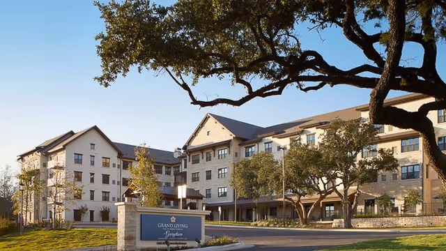 Front exterior of the multi-story Grand Living at Georgetown senior living building with trees and a sign in the foreground.