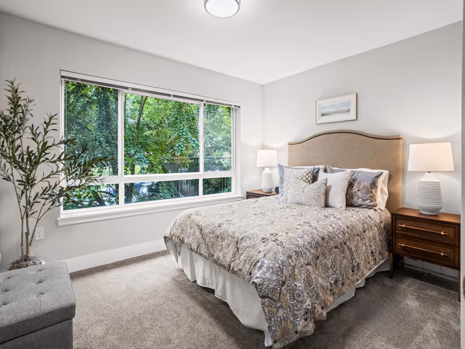 A bright bedroom with a large window showing green trees outside. The room features a bed with a beige upholstered headboard, patterned bedding, and multiple pillows. There are two wooden nightstands on either side of the bed, each with a white table lamp. A framed picture hangs above the bed, and a potted plant is placed near the window. The floor is carpeted in a neutral color.