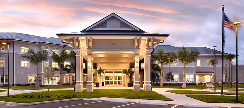 Front exterior view of The Preserve in Fort Myers senior living facility at dusk, featuring a large covered entrance with columns, palm trees, well-maintained landscaping, and an American flag on a flagpole.