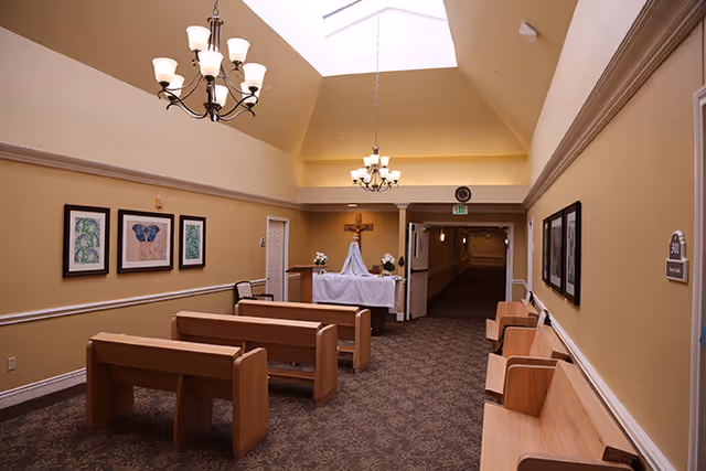 Interior view of a small chapel or prayer room with wooden pews, a table covered with a white cloth holding a crucifix and flowers, beige walls, framed artwork, carpeted floor, and two chandeliers hanging from a high ceiling with a skylight.