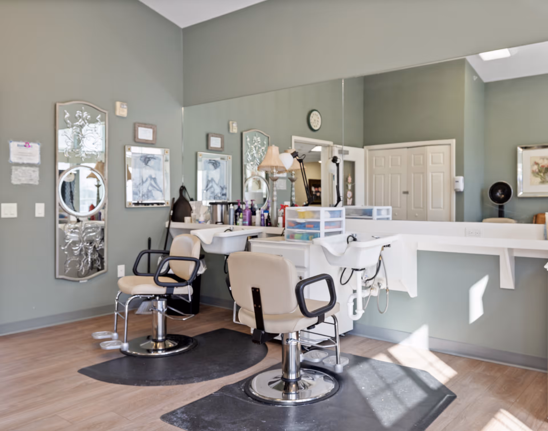 Interior view of a salon area in a senior living facility with two beige salon chairs in front of a large mirror, a white sink for hair washing, various hair care products on the counter, and decorative wall mirrors on a light green wall.