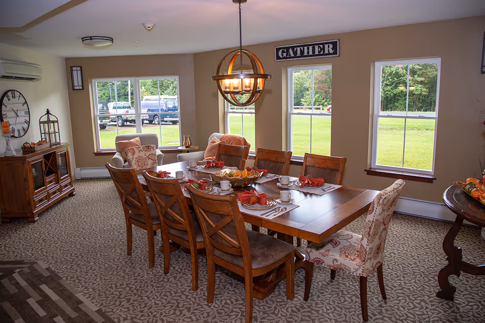 A dining room with a wooden table set for six people with plates, cups, and red napkins. The room has three large windows showing a green outdoor area. There is a decorative sign above the windows that says 'GATHER'. The room features patterned carpet, a wooden sideboard with decorative items, and two armchairs near the windows.