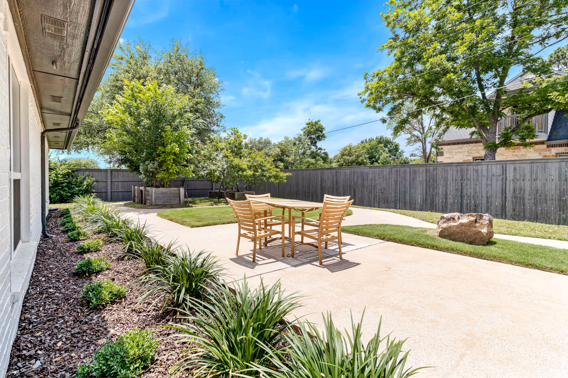 Outdoor patio area with a wooden table and four chairs on a concrete surface, surrounded by green plants, trees, and a wooden fence under a clear blue sky.