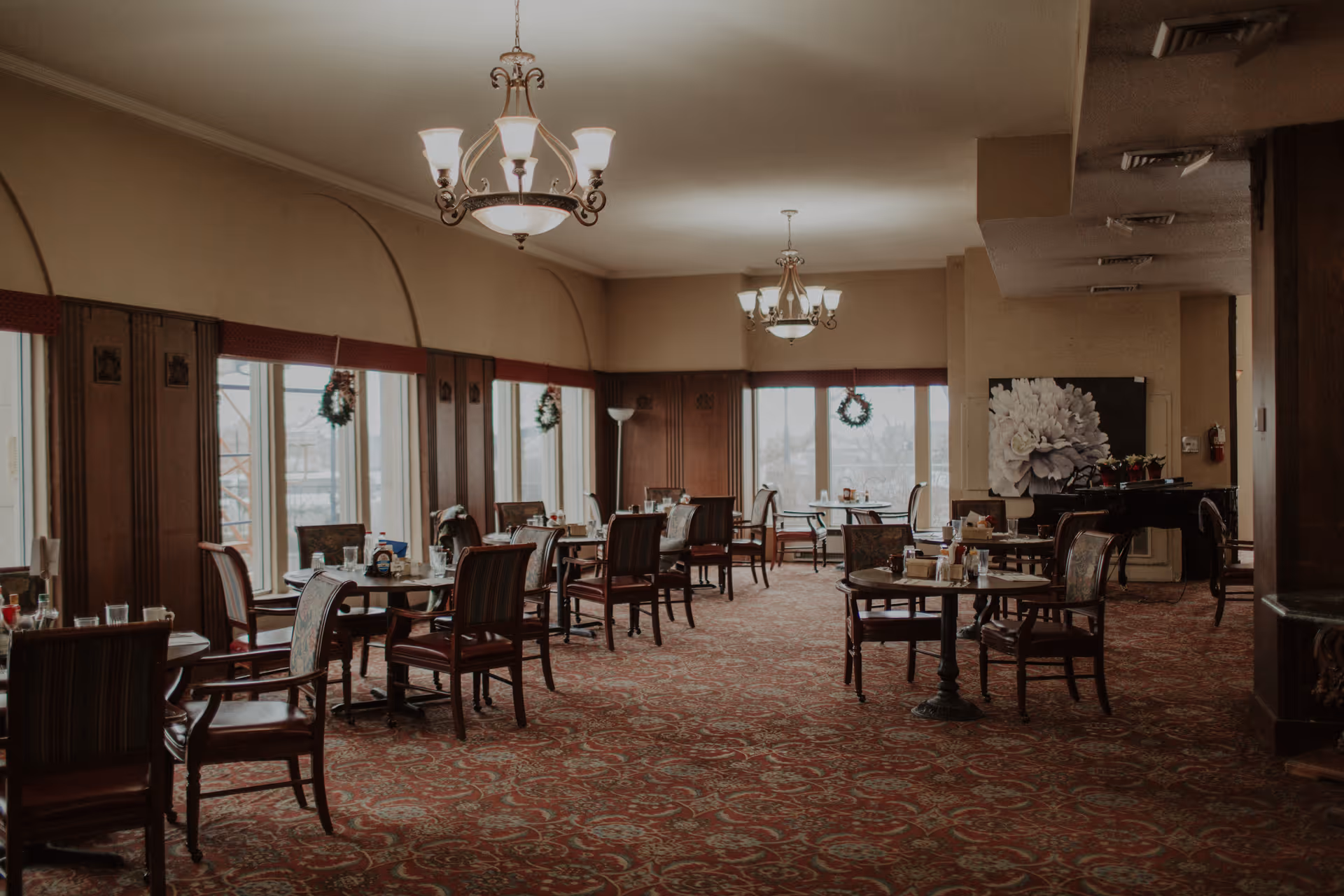 A spacious dining room with multiple round tables and chairs arranged neatly. The room features large windows decorated with wreaths, allowing natural light to enter. The carpet has an intricate pattern, and elegant chandeliers hang from the ceiling. There is a large floral artwork on one wall and a piano in the corner.