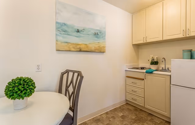Small kitchen area with beige cabinets, a white refrigerator, a sink, and a countertop. A round white table with a green potted plant and a wooden chair are visible. A painting of a beach scene hangs on the wall.