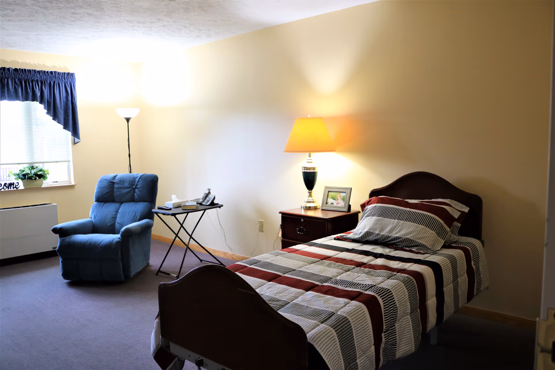 A cozy bedroom with a single bed covered in a striped comforter in shades of red, white, and black. Next to the bed is a wooden nightstand with a lamp and a framed photo. A blue recliner chair is positioned near a window with blue curtains, and a small folding table holds a telephone and a box of tissues. The room has beige walls and carpeted floor.
