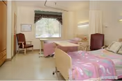 A bright and clean room with two hospital-style beds, each with pink bedding. There is a window with a valance letting in natural light, a wooden chair near the window, and a small bedside table between the beds. The walls are light-colored, and the floor is a light tile.