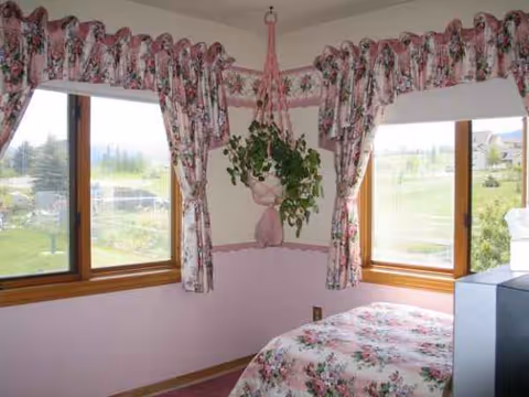 Pastel pink bedroom with floral quilt, hanging plant, and large windows with floral curtains.