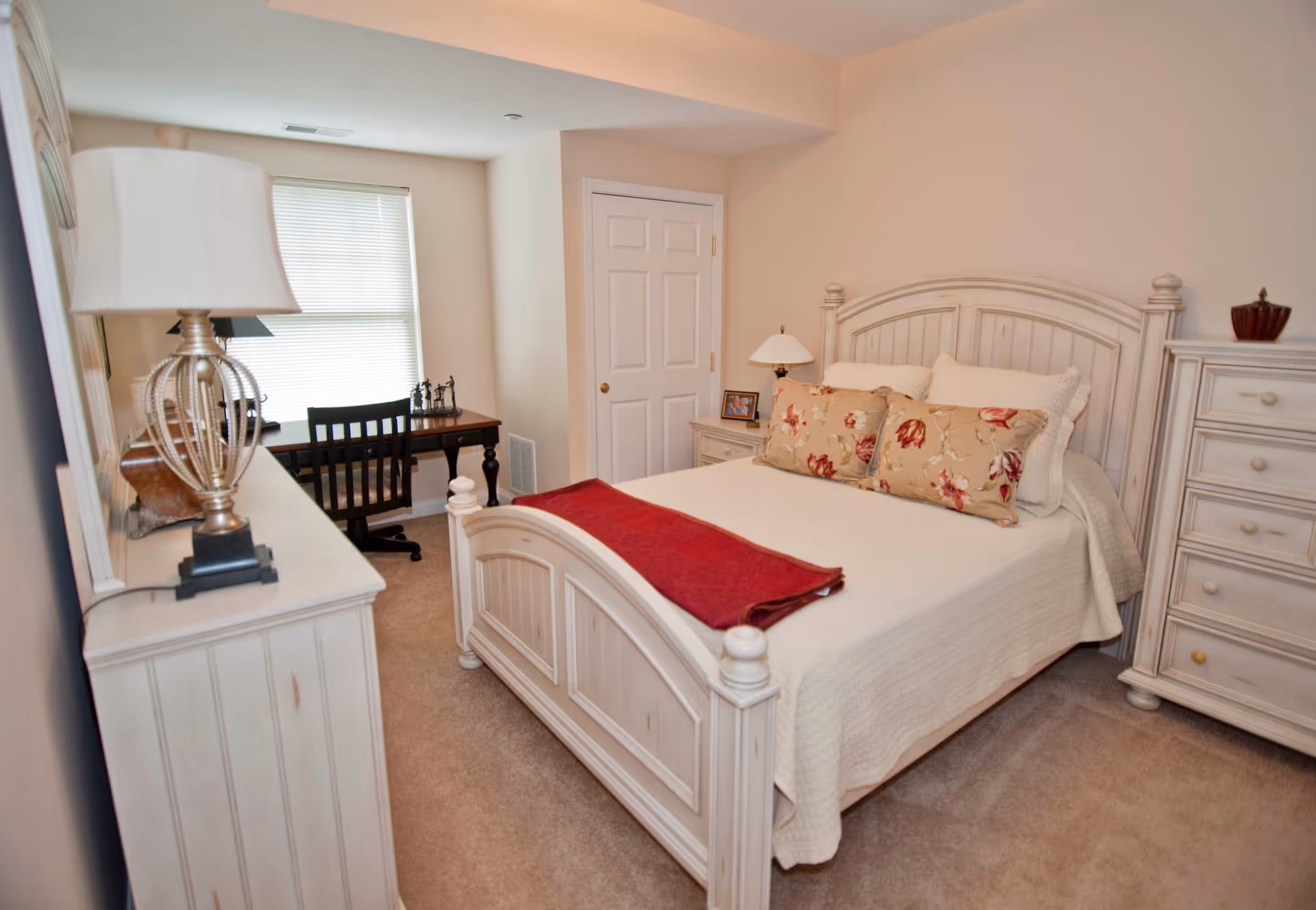 A cozy bedroom with a white wooden bed frame, a bed made with white bedding and floral pillows, a red throw blanket at the foot of the bed, a matching white wooden dresser and chest of drawers, a bedside table with a lamp and a framed photo, and a small desk with a chair near a window with blinds.