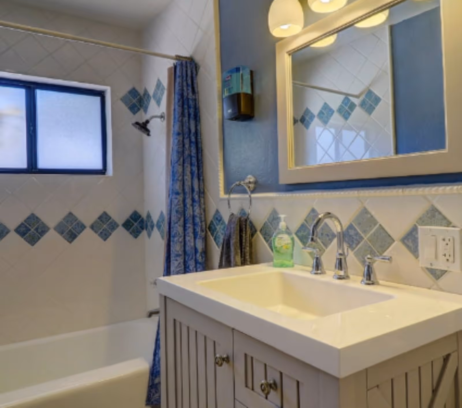 Bright bathroom with a white sink vanity and mirror, tiled walls with blue diamond accents, and a bathtub with a blue patterned shower curtain.