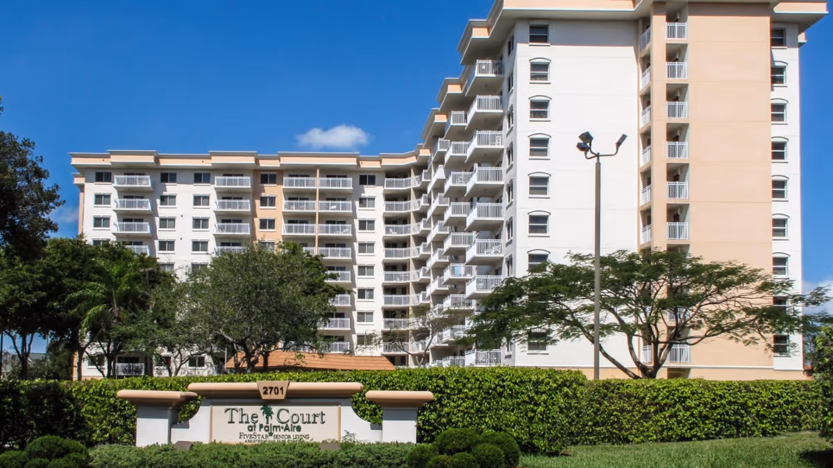 Multi-story senior living building with balconies and a front sign reading 'The Court at Palm Aire' behind manicured hedges and trees under a blue sky.