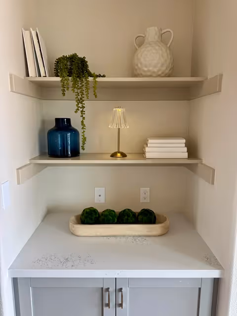 A kitchen nook with shelves displaying decor and a tray of green fruit on the counter.
