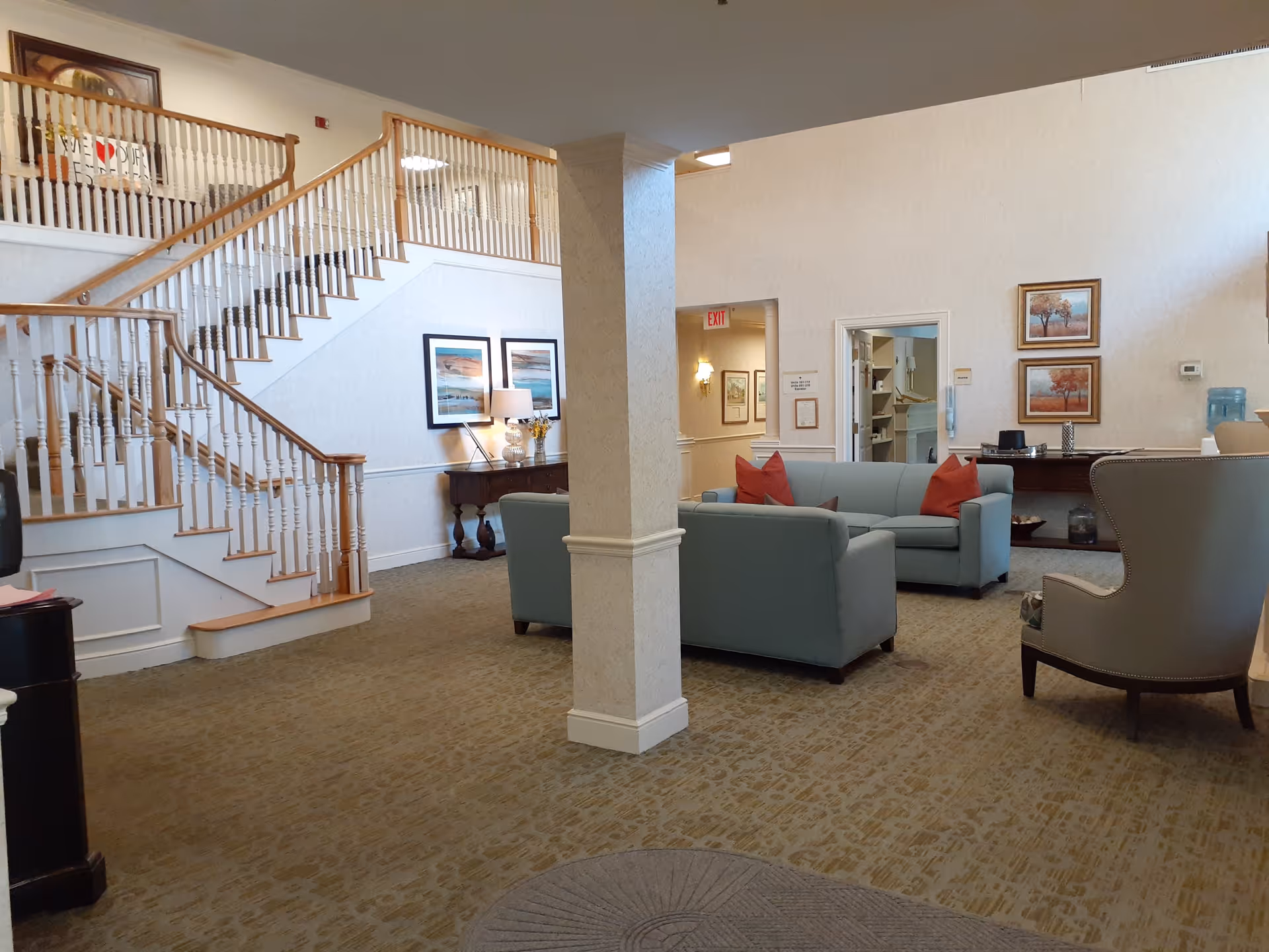 Interior view of a senior living facility lounge area featuring a staircase with wooden railings, light blue sofas with red pillows, a beige armchair, framed artwork on the walls, a table with a lamp, and a water cooler in the background.