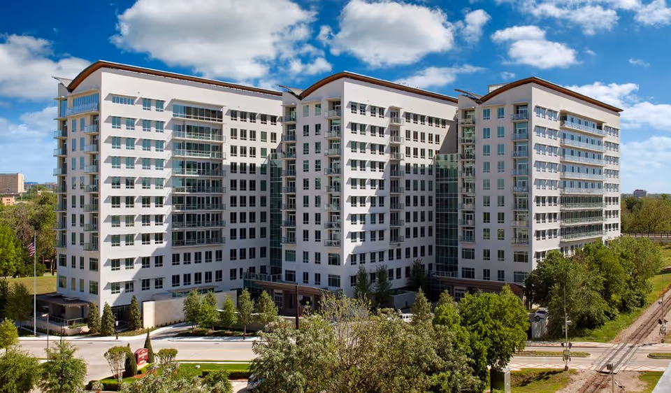 A large modern multi-story senior living facility building with white exterior walls, numerous windows, and balconies. The building is surrounded by green trees and landscaping under a partly cloudy blue sky.