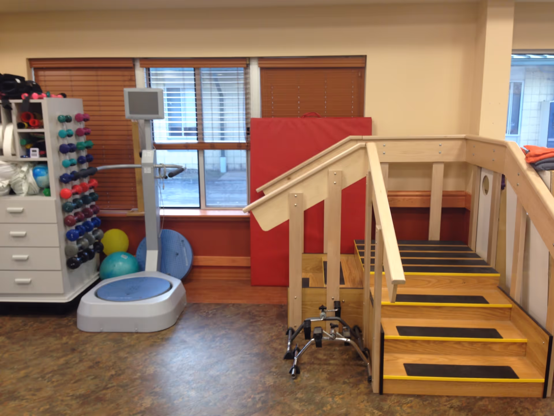 A physical therapy or rehabilitation room featuring a set of wooden stairs with handrails, a walker, a balance platform machine, various colorful dumbbells on a rack, exercise balls, and a red padded mat against the wall. The room has windows with wooden blinds and a brown floor.