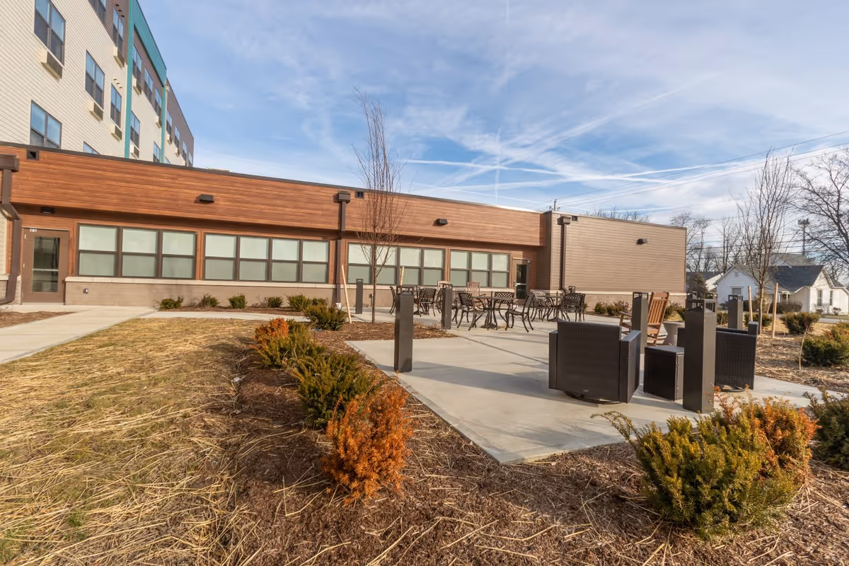 Outdoor patio area at a senior living facility with several tables and chairs arranged on a concrete surface. The building exterior features large windows and wood paneling. There are small bushes and young trees planted around the patio, with a clear sky overhead.