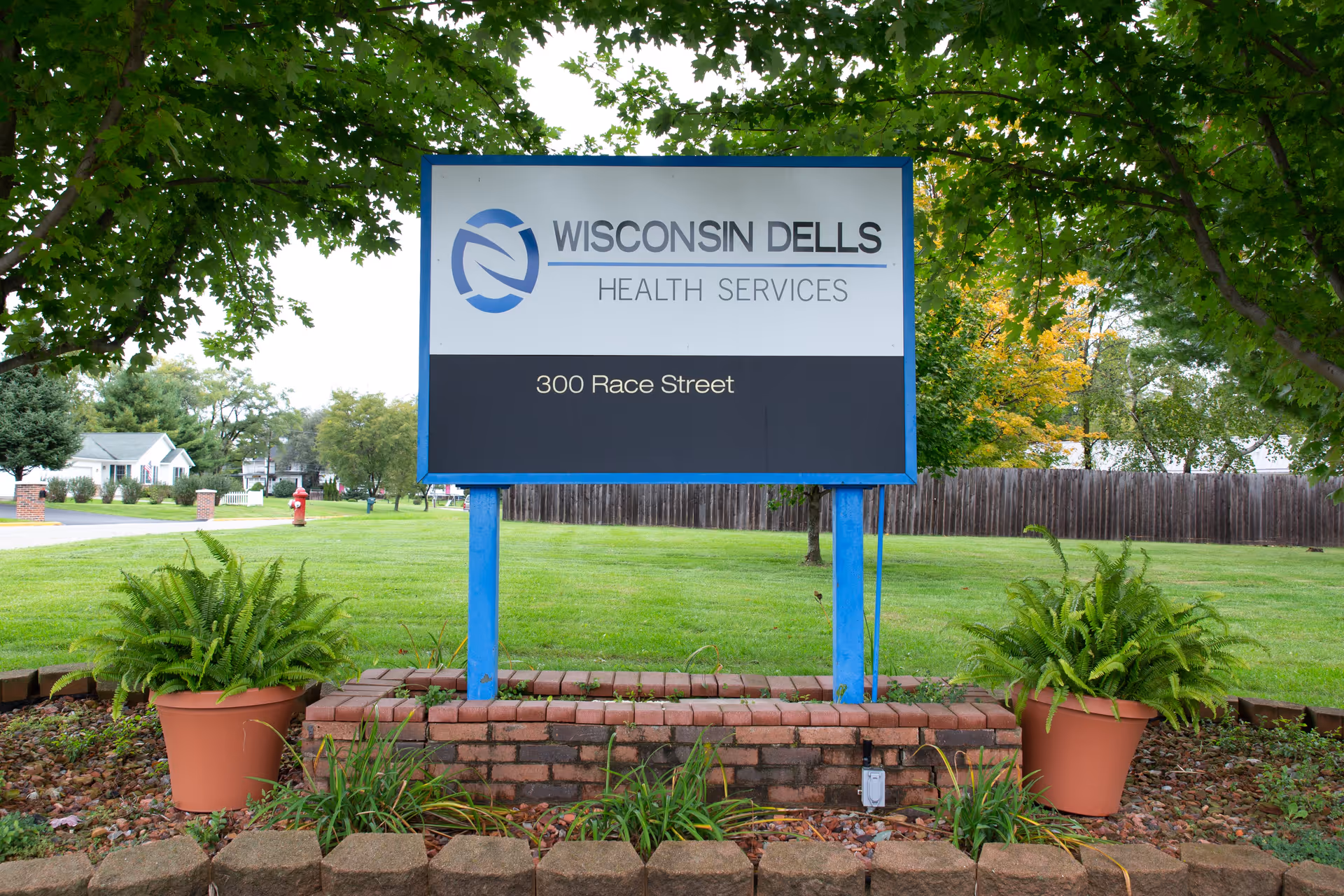 Outdoor view of a sign for Wisconsin Dells Health Services located at 300 Race Street, surrounded by green grass, trees, and two potted plants on either side of the sign.