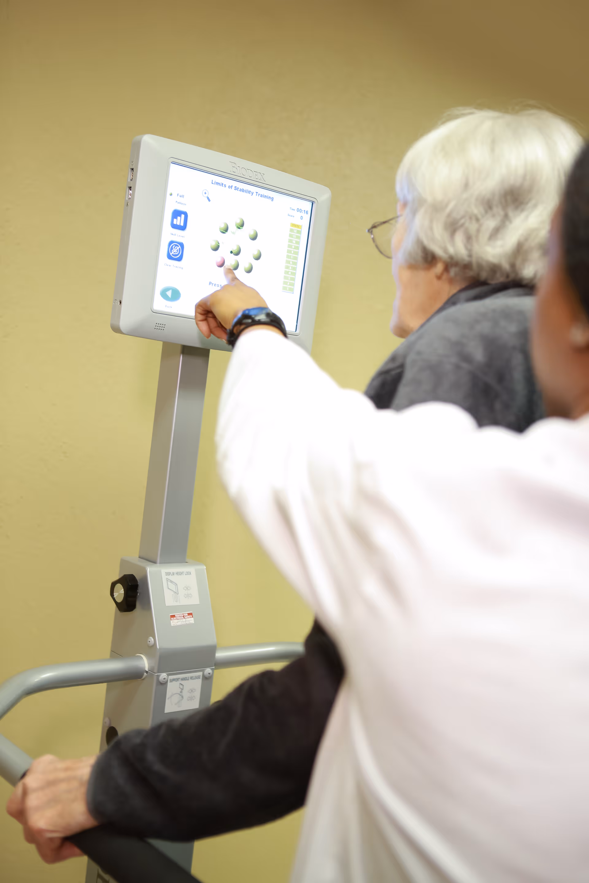 An elderly person with white hair and glasses is using a stability training machine with a digital screen, while another person points at the screen to assist.