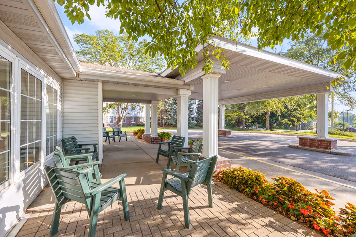 Outdoor covered seating area with green chairs arranged on a paved patio next to a building with large windows. There are trees providing shade and flower beds with colorful flowers along the edge of the patio. The area opens to a driveway or parking lot with more greenery in the background.
