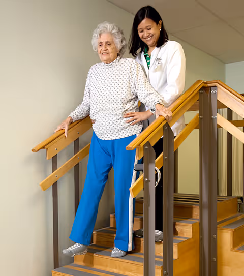 An elderly woman wearing a white patterned sweater and blue pants is walking down a wooden staircase with handrails on both sides, assisted by a smiling female caregiver dressed in a white coat.