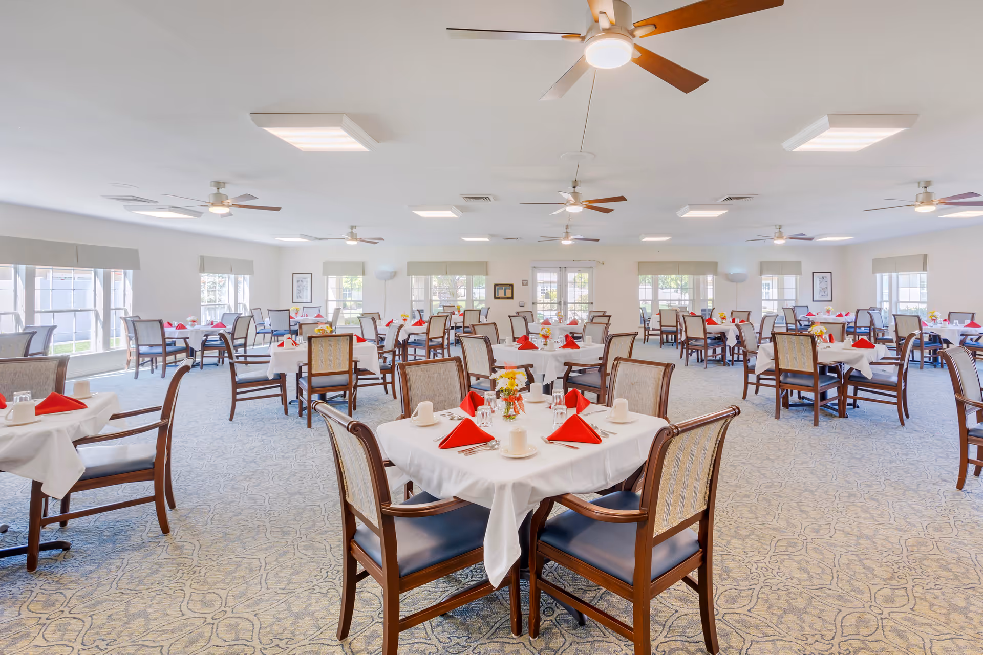 A spacious dining room with multiple tables covered in white tablecloths, each set with red folded napkins, cups, glasses, and silverware. The room has large windows allowing natural light to fill the space, ceiling fans, and light fixtures on the ceiling. The carpet has a subtle pattern and the chairs have wooden frames with cushioned seats and backs.