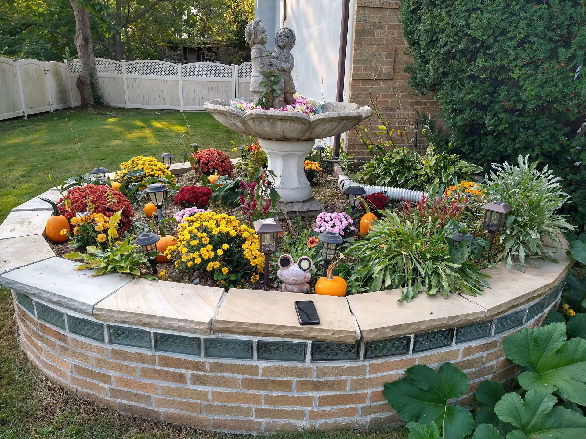 Raised circular brick planter in a backyard containing a decorative birdbath, colorful flowers, small pumpkins and solar garden lights.