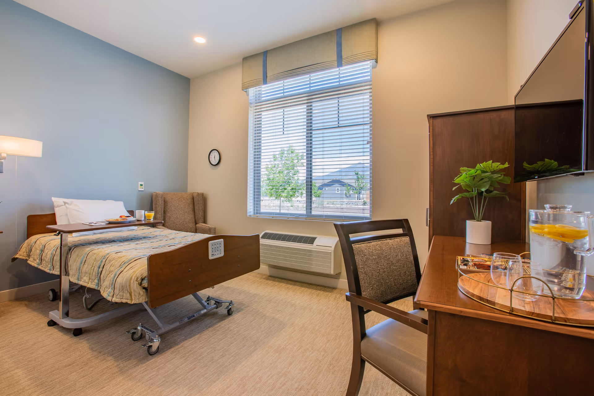 A bright and clean patient room in a transitional care facility featuring a hospital bed with a meal tray, a comfortable armchair, a window with blinds showing an outdoor view, a desk with a chair, a potted plant, and a pitcher of water with lemon slices.