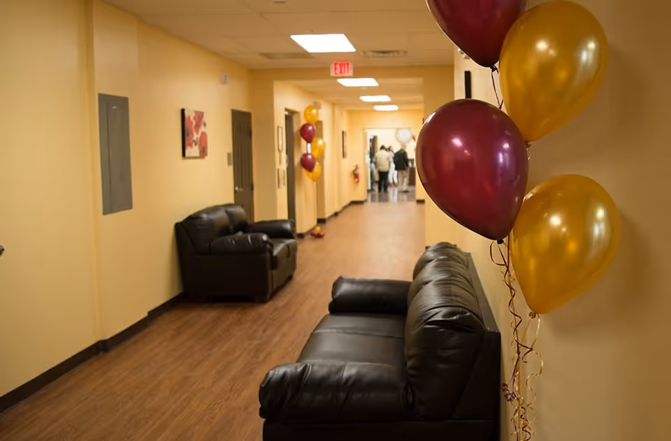 A hallway in a senior living facility with beige walls and wood flooring. There are two black leather couches along the walls and clusters of maroon and gold balloons tied to the wall. At the far end of the hallway, a group of people is gathered near a doorway. The hallway is well-lit with ceiling lights and has a fire extinguisher mounted on the wall.