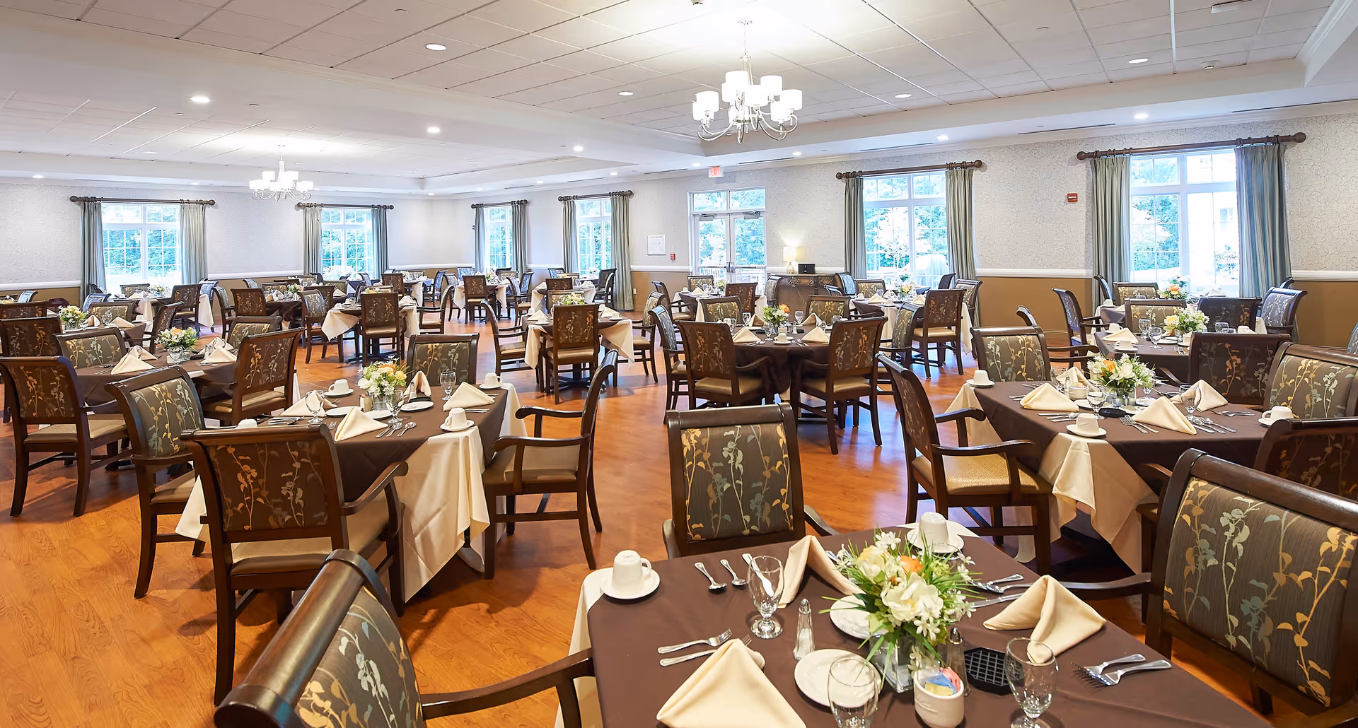 A spacious dining room with multiple tables covered in brown and beige tablecloths, each set with napkins, cups, glasses, and floral centerpieces. The room has wooden flooring, large windows with curtains, and ceiling lights providing bright illumination.