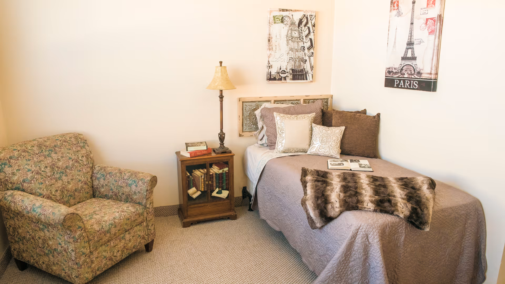 A cozy bedroom with a single bed covered in a taupe quilt and multiple decorative pillows. A faux fur throw is draped over the foot of the bed. Next to the bed is a wooden nightstand with a glass door containing books, topped with a table lamp and two books. To the left is a floral upholstered armchair. The walls are decorated with two framed pictures, one featuring the Eiffel Tower and the other a vintage London scene.
