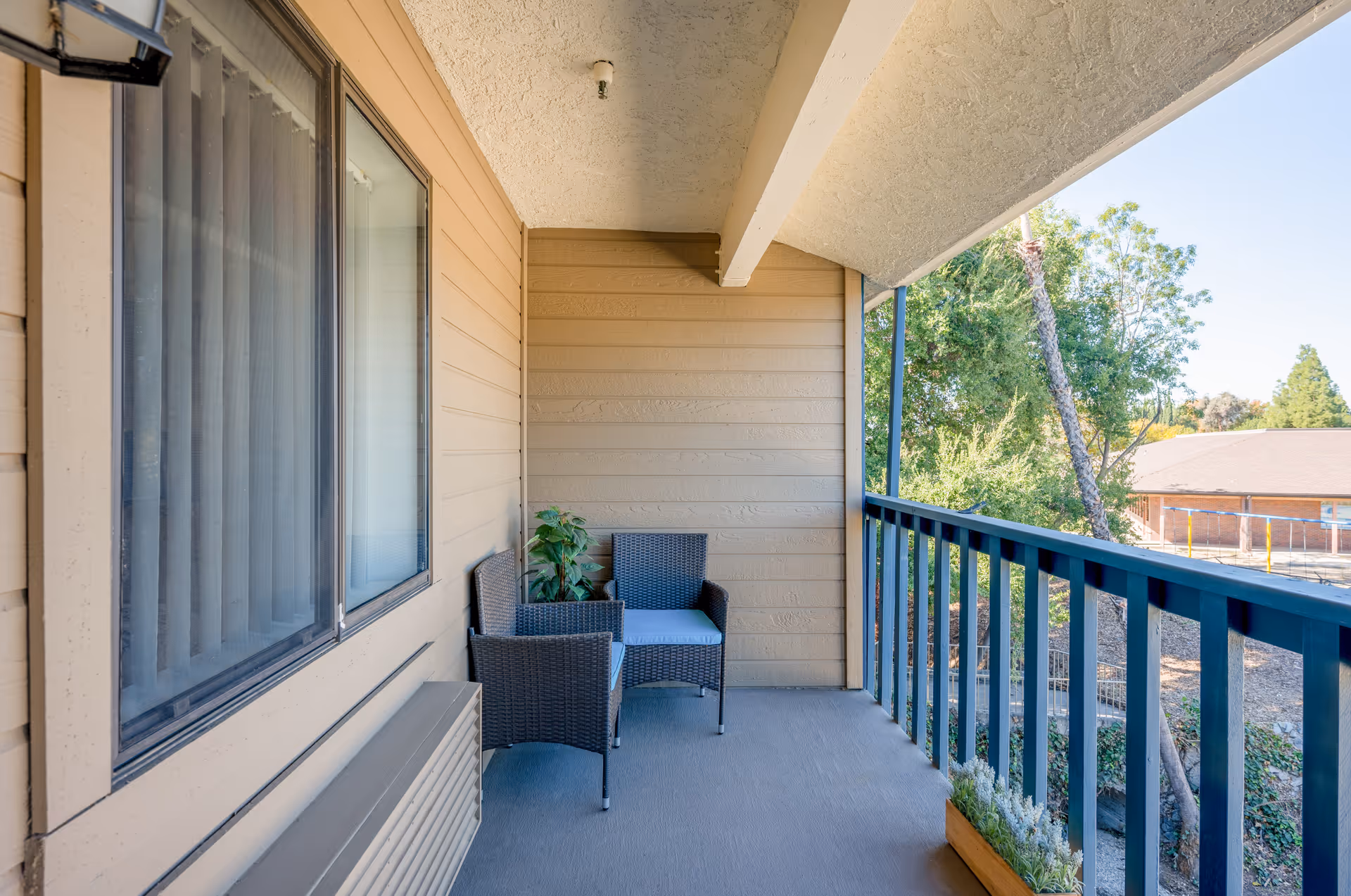 A covered balcony with two wicker chairs with cushions, a small plant in the corner, and a planter box with greenery along the railing. The balcony overlooks trees and a building in the distance under a clear blue sky.