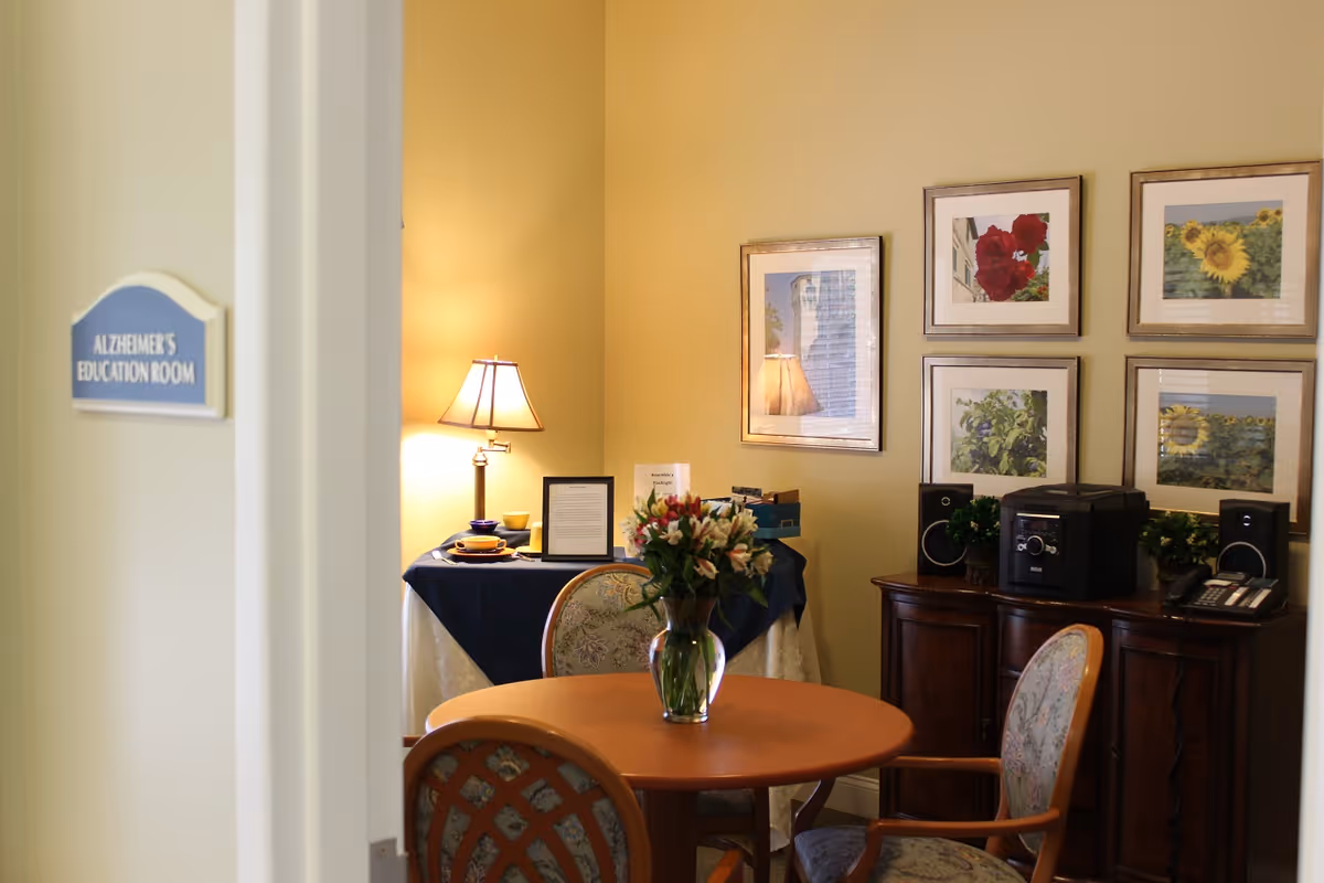 Small furnished interior room with a round table and chairs, a vase of flowers, framed artwork, and a door sign that reads 'ALZHEIMER'S EDUCATION ROOM'.