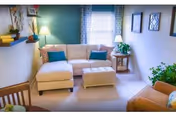 A cozy living room with a beige sectional sofa adorned with colorful throw pillows, a matching ottoman, a wooden side table with a lamp, framed artwork on the walls, a wooden chair, a small shelf with books and decor, and a window with patterned curtains letting in natural light.