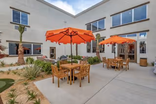 Outdoor courtyard area at Palm Vista Senior Living with wooden tables and chairs shaded by bright orange umbrellas, surrounded by desert landscaping and a modern building with large windows.