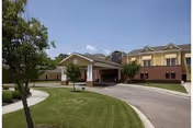 Front exterior of a two-story senior living facility with a covered entrance, circular driveway, and landscaped lawn under a blue sky.
