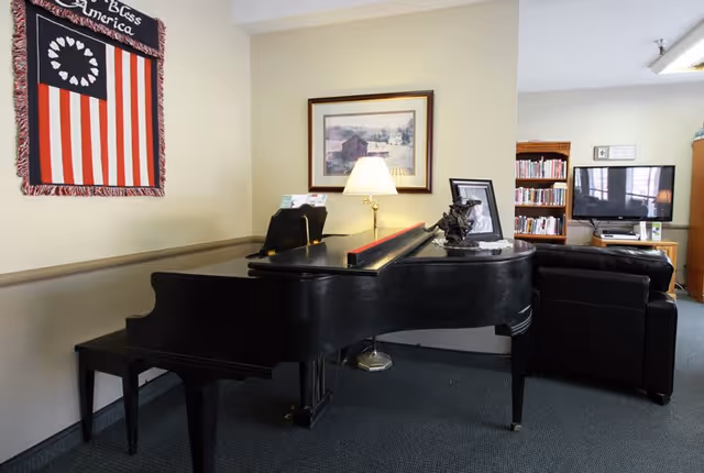 Interior room with a black grand piano in the foreground, a bench, a table lamp on the piano, a framed picture on the wall, a patriotic wall hanging with the text 'God Bless America', a bookshelf filled with books, a flat-screen TV, and a black leather armchair.
