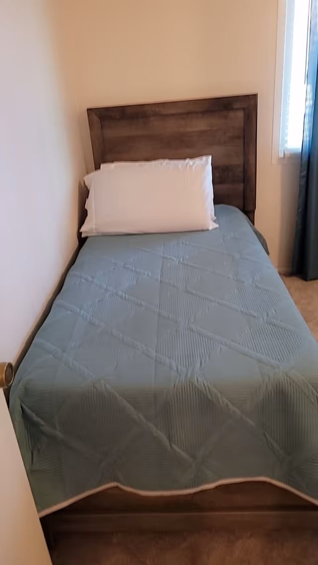 A single bed with a wooden headboard, covered with a blue quilted bedspread and a white pillow, placed against a beige wall next to a window with blue curtains.