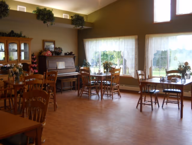Sunlit dining room with wooden tables and chairs, a piano against the wall, and large windows with lace curtains.