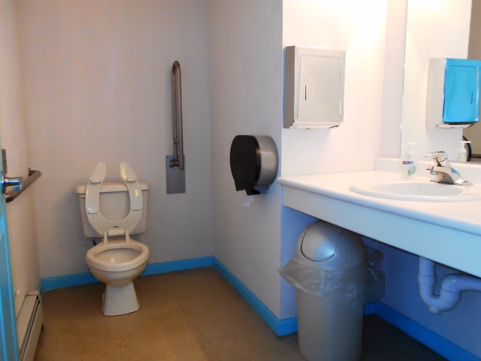 A clean, accessible bathroom with a toilet featuring an open seat, a grab bar on the wall, a sink with a faucet, a wall-mounted paper towel dispenser, a toilet paper dispenser, and a trash can underneath the sink.