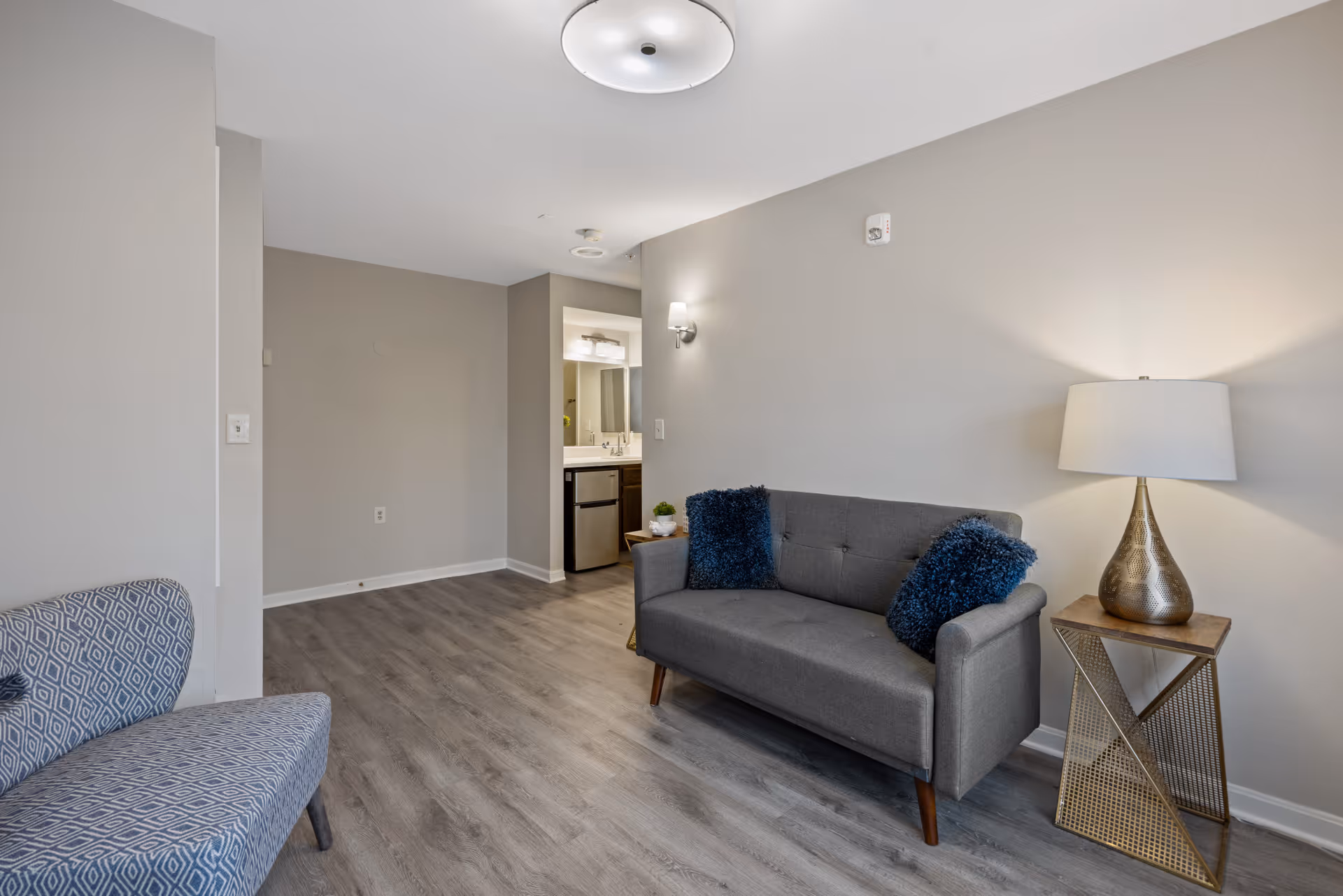 A modern living room area with a gray sofa adorned with two dark blue pillows, a patterned armchair, a wooden side table with a decorative lamp, and light gray walls and flooring. In the background, there is a small kitchenette area with a sink, mirror, and mini refrigerator.