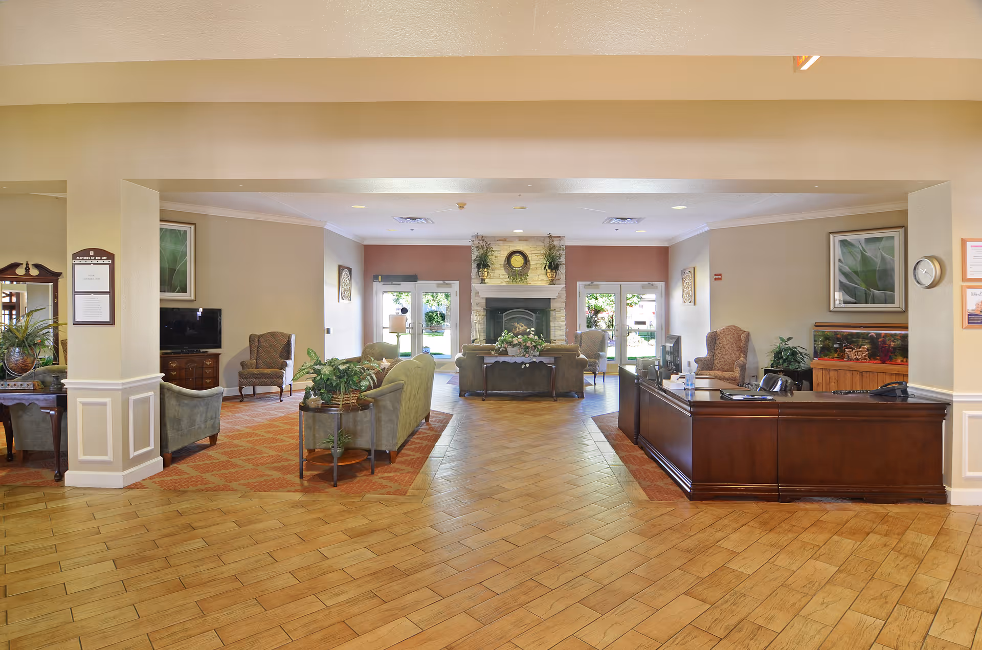 A spacious and well-lit senior living facility common area with a wooden reception desk on the right, comfortable seating including armchairs and sofas arranged around a central fireplace, decorative plants, framed artwork on the walls, and large glass doors letting in natural light.