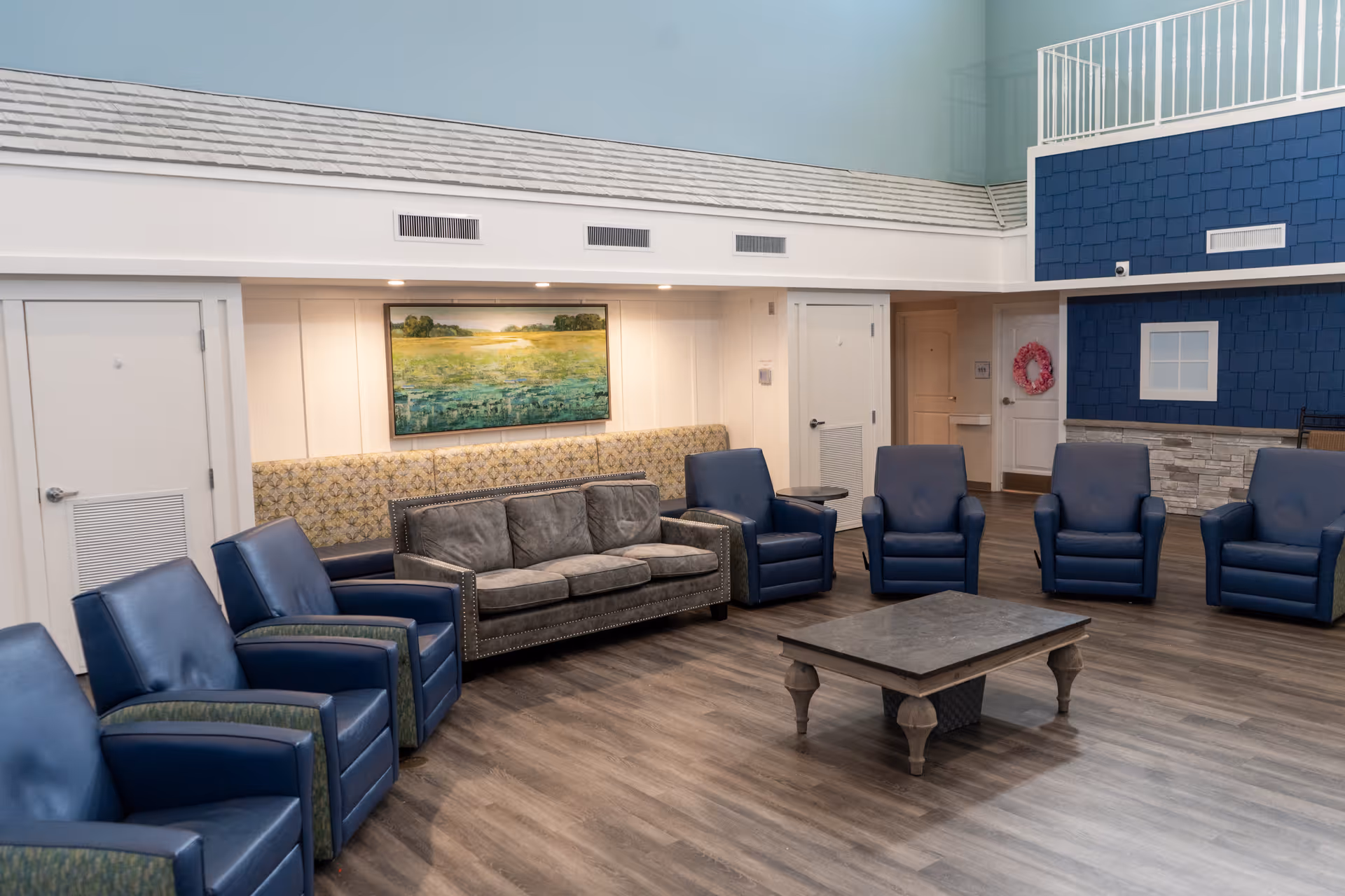 A spacious seating area with multiple blue armchairs and a gray sofa arranged around a wooden coffee table on a wood floor. The walls are light-colored with a large landscape painting and a decorative blue section resembling exterior siding. There are doors and a small table in the background.