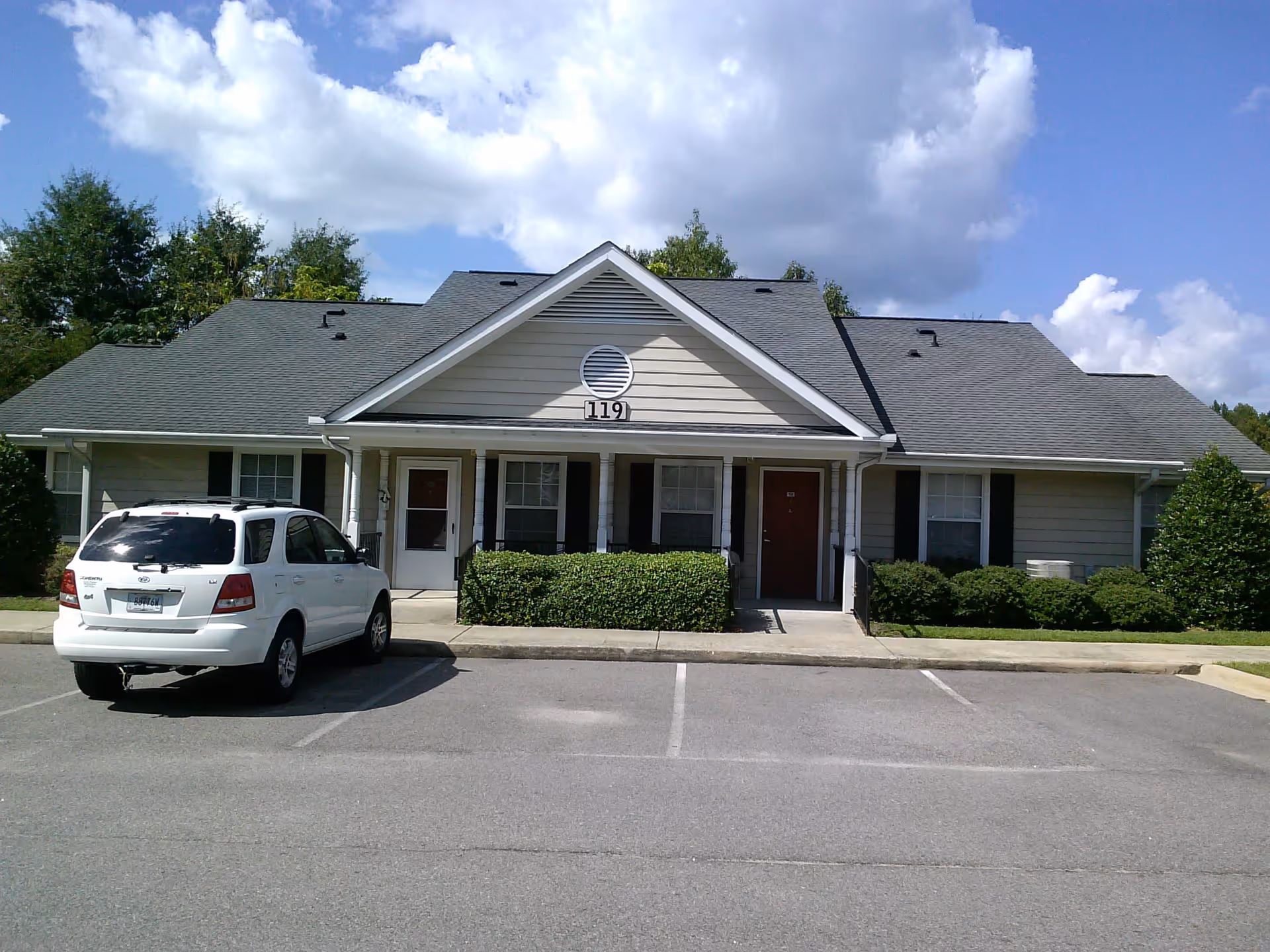Exterior view of a single-story residential building with two front doors, numbered 119, surrounded by bushes and trees. A white SUV is parked in front of the building under a partly cloudy sky.
