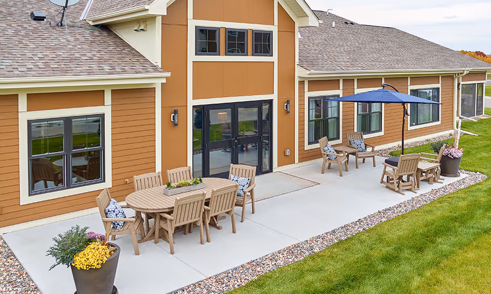 Outdoor patio area at The Lakes at Stillwater featuring a concrete seating area with wooden tables and chairs, some with blue patterned cushions. There is a large blue umbrella providing shade over one seating area. The patio is bordered by decorative rocks and green grass, with the building's brown exterior and multiple windows in the background.