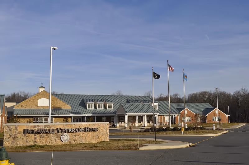 Front exterior of the Delaware Veterans Home with a stone sign, flagpoles, and the building entrance under a clear blue sky.