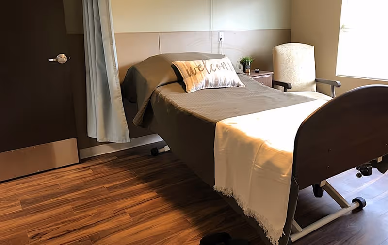 A single bed with a brown blanket and a white throw blanket at the foot, featuring a decorative pillow with the word 'welcome'. The bed is positioned next to a window with natural light coming in. There is a beige armchair and a small wooden nightstand with a plant and other small items. The floor is wooden, and a door with a metal kick plate is visible on the left side.