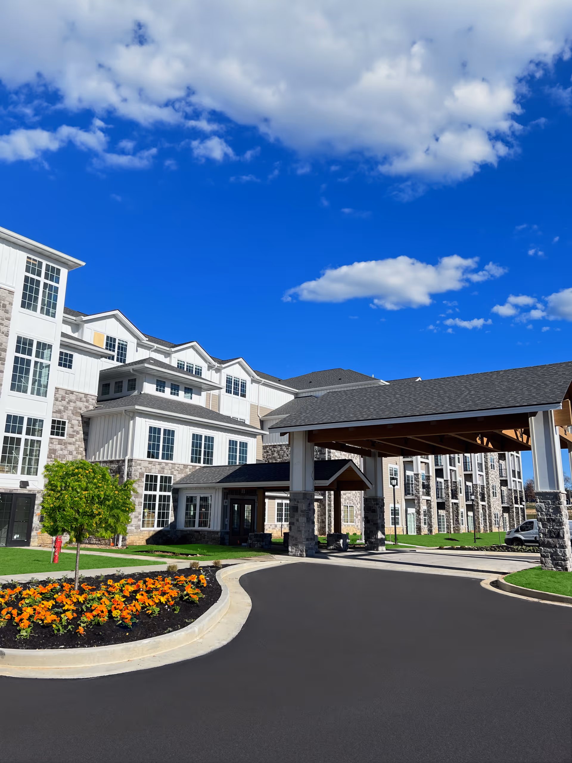 Covered entrance and porte-cochère of a multi-story senior living building with landscaped flower beds and a bright blue sky.