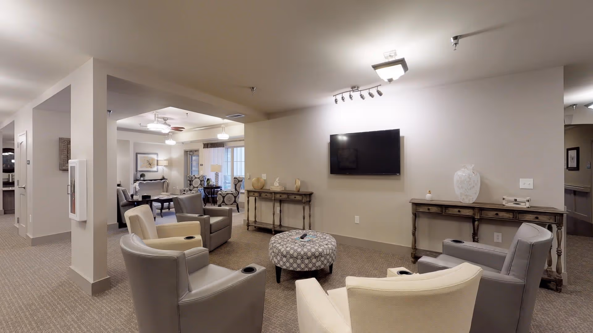 A cozy living room area in a senior living facility with four armchairs arranged around a patterned ottoman. A flat-screen TV is mounted on the wall above two wooden console tables decorated with vases and small items. The room has neutral-colored walls and carpeted floors, with additional seating and a dining area visible in the background.