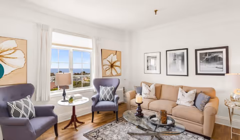 A bright and cozy living room with a beige sofa adorned with decorative pillows, two purple armchairs with patterned cushions, a glass coffee table on a patterned rug, and wall art including two large floral paintings and three black-and-white framed photographs. A window with white curtains offers a view of the outdoors.