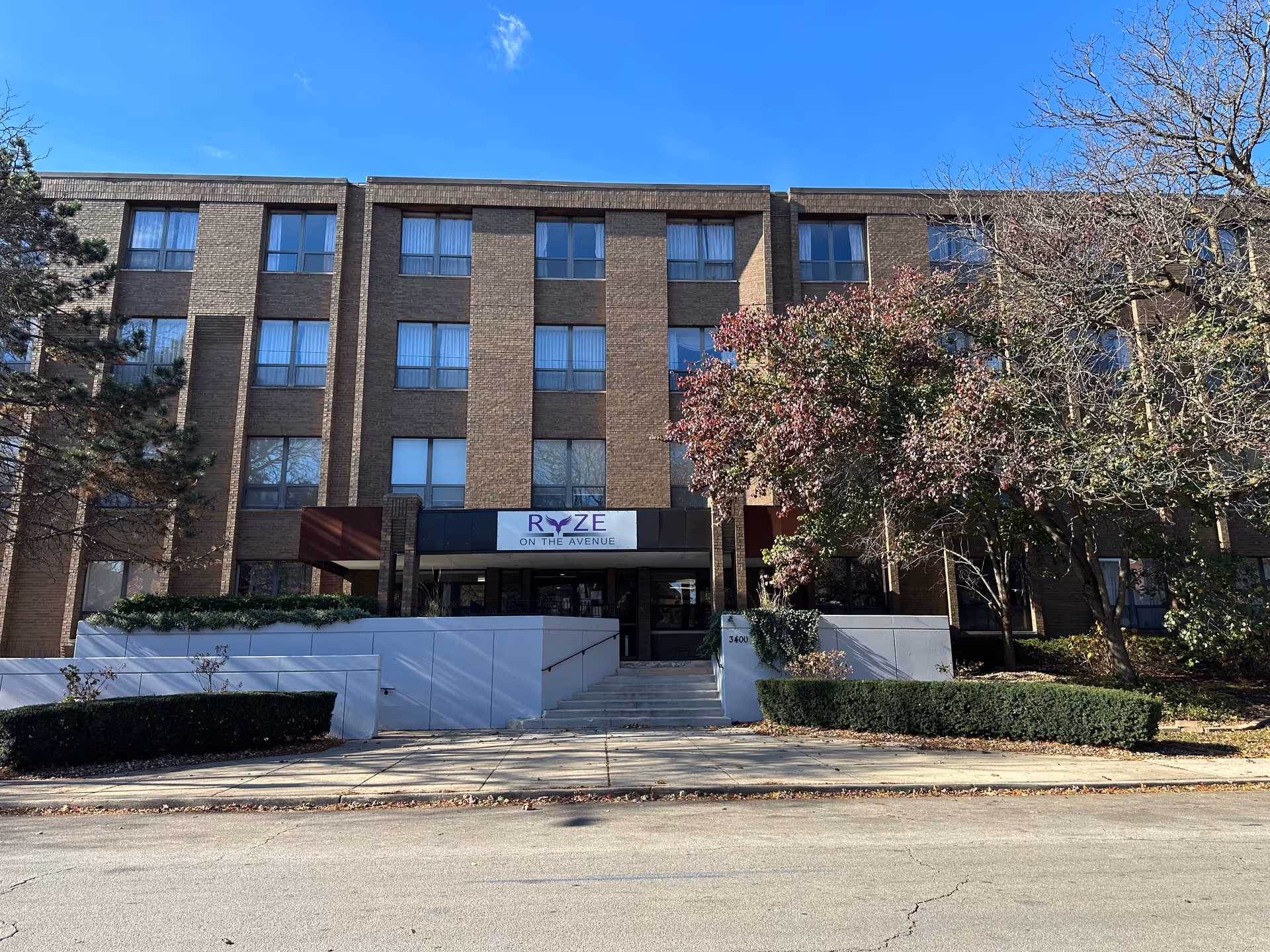 Front exterior of a four-story brick building with a 'RYZE on the Avenue' sign above the main entrance and trees flanking the walkway.